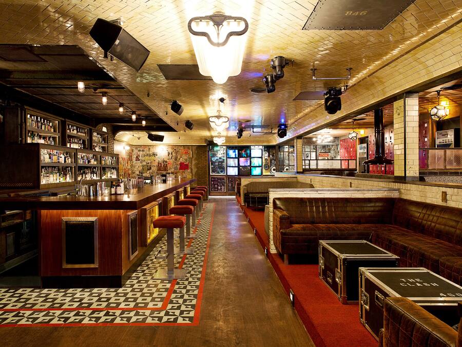 Uma área de bar com um balcão comprido, bancos e piso estampado. Há também cabines de couro e um espaço de bar com prateleiras cheias de garrafas.