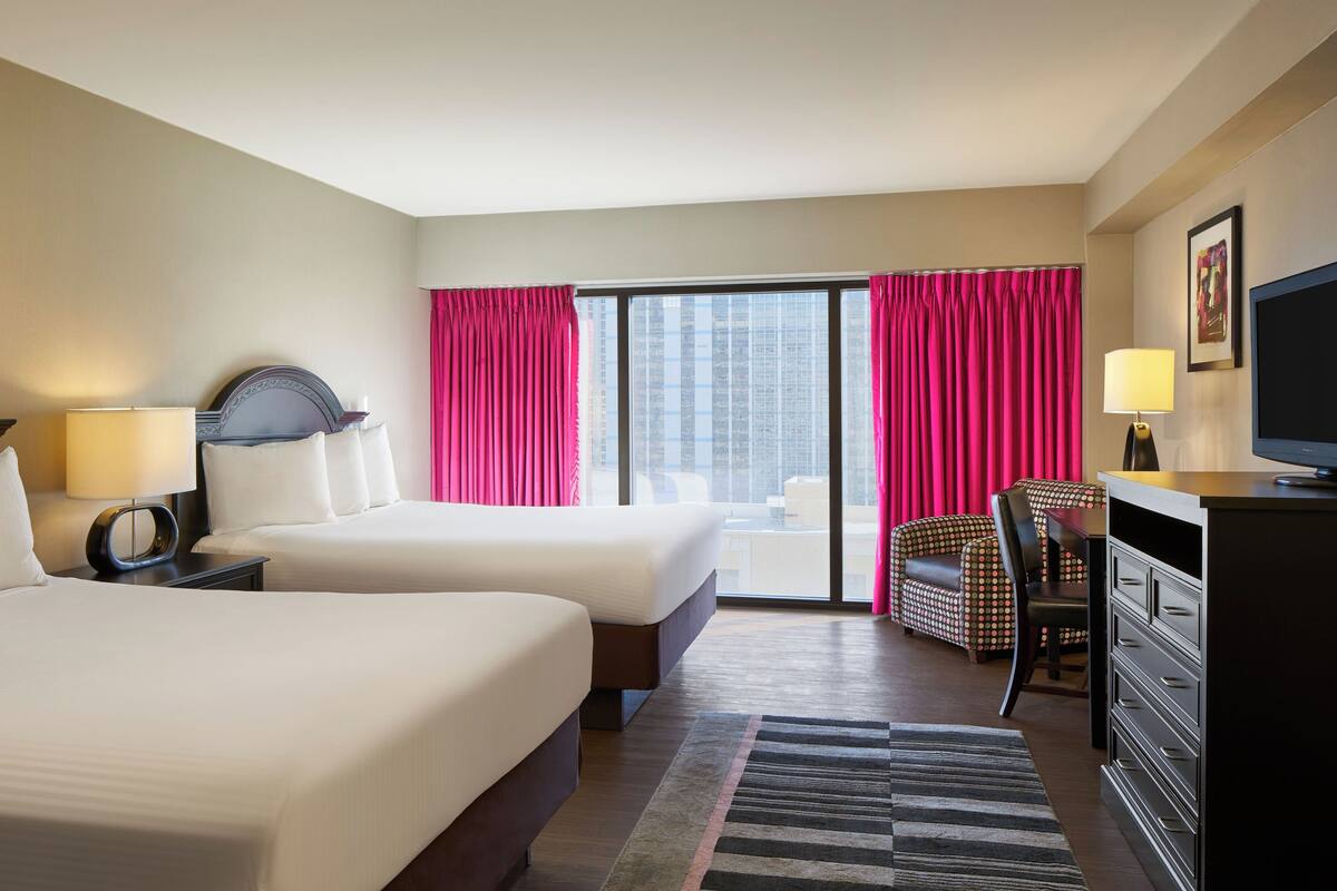 A hotel room with two beds, a desk with a television, and a view of the city through large windows.