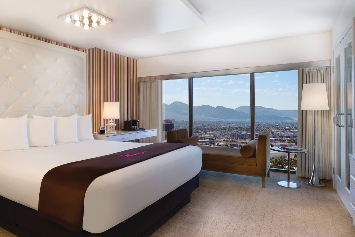 A hotel room with a large bed, a desk, a chair, and a view of a cityscape and mountains.
