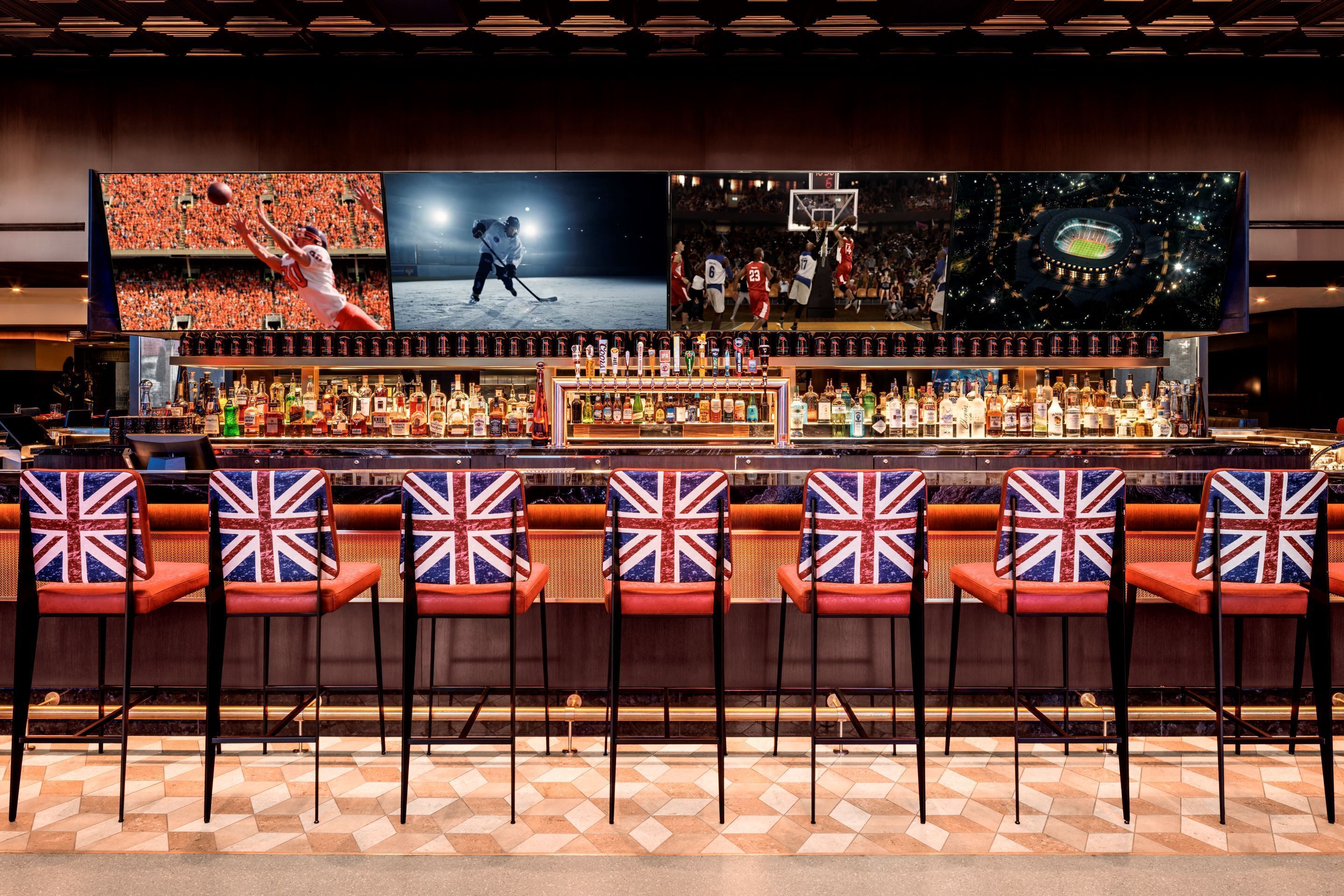 A bar with multiple screens displaying sports events, a variety of liquor bottles, and bar stools with Union Jack designs.