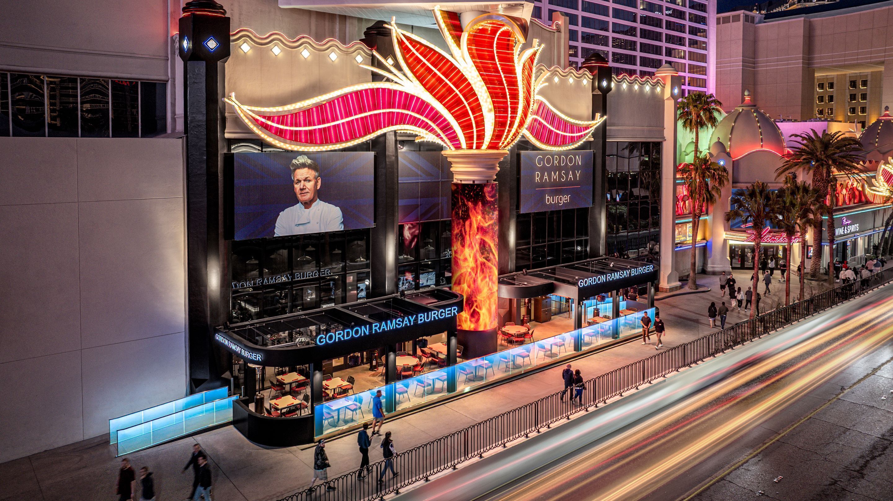 Ein Gordon Ramsay Burger Restaurant mit einer großen, beleuchteten Schautafel, Außenbereich und Blick auf die Straße.