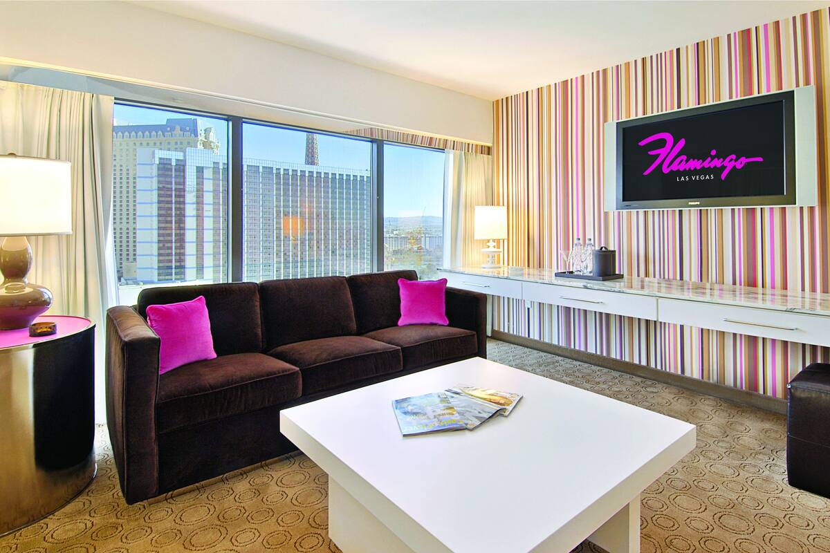 A hotel room with a large window, a sofa with pink cushions, a white coffee table, and a TV on the wall displaying "Flamingo Las Vegas".