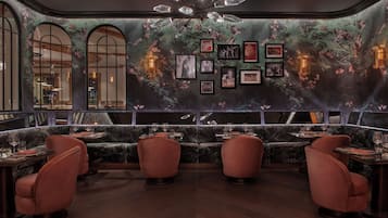 A dimly lit restaurant with a dark, floral wallpaper, red upholstered chairs, and framed pictures on the wall.
