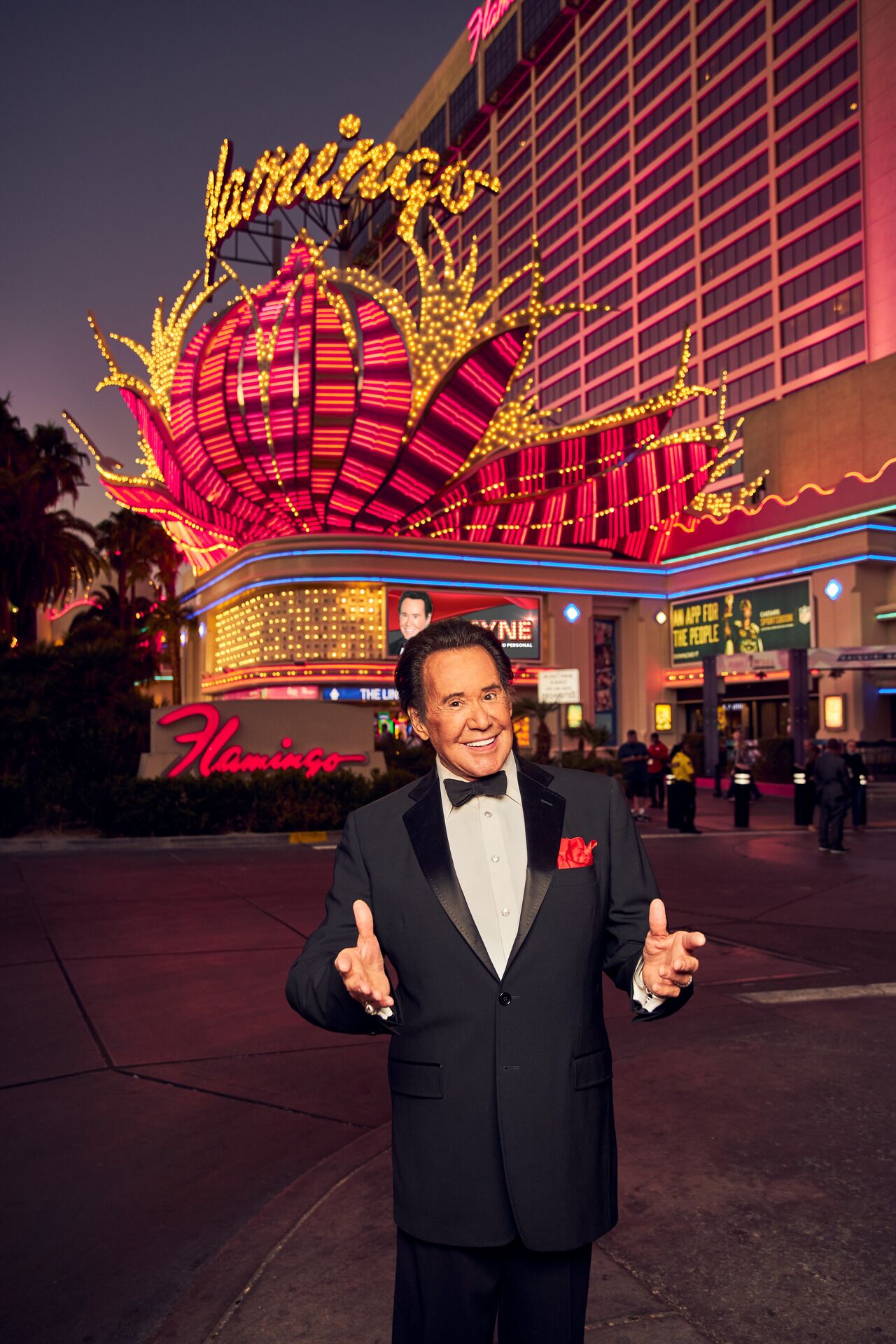 A man in a black suit with a red rose boutonniere standing in front of a brightly lit building with a large, illuminated sign that reads "Hammond's".