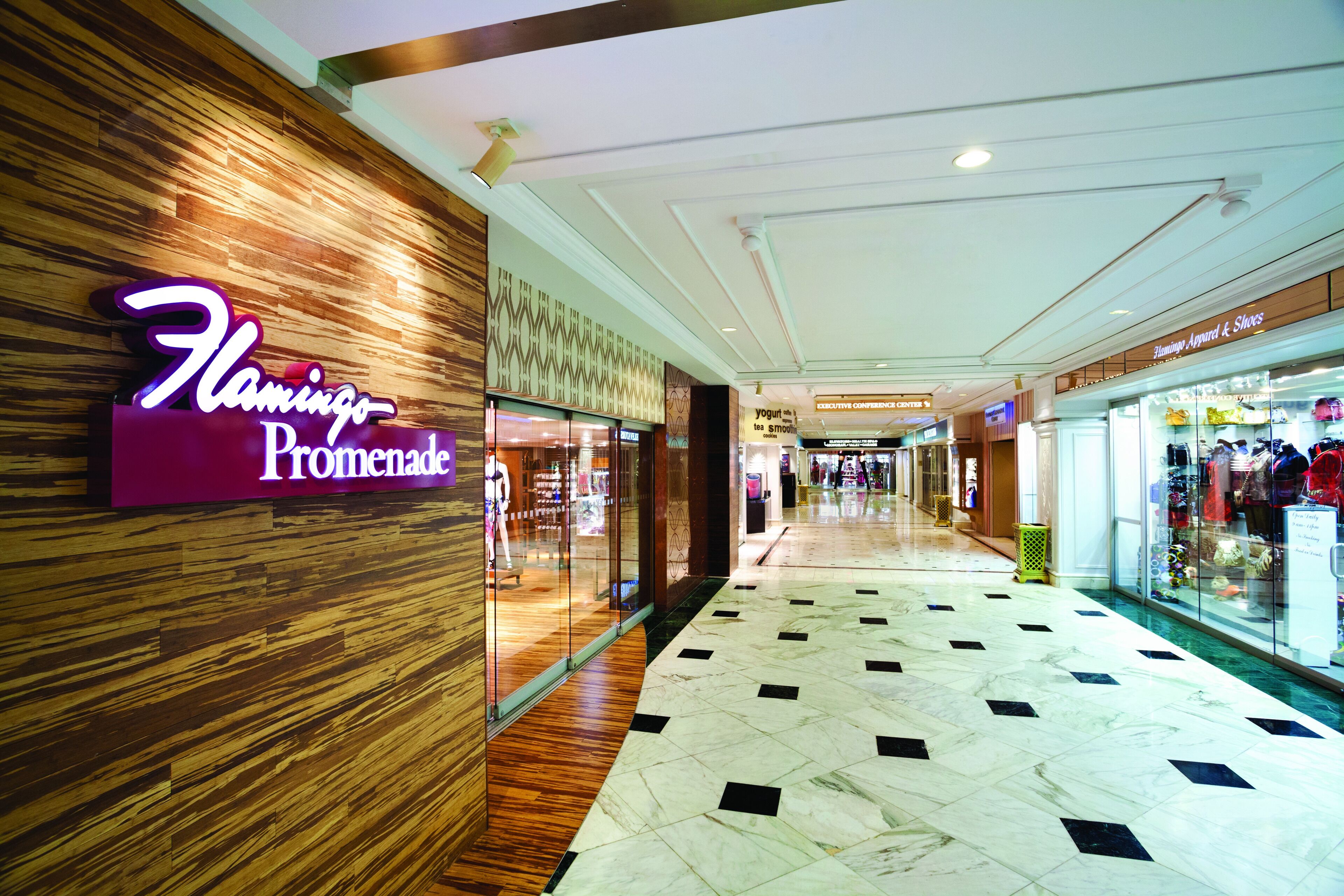 Um corredor de shopping com um letreiro de neon para Flamingo Promenade, uma loja chamada Executive Conference Center e uma loja de sapatos.