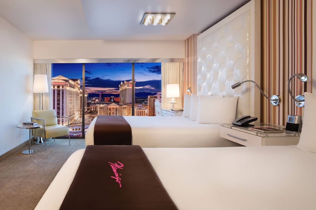 A hotel room with a large bed, a desk, a chair, and a view of a cityscape at night.
