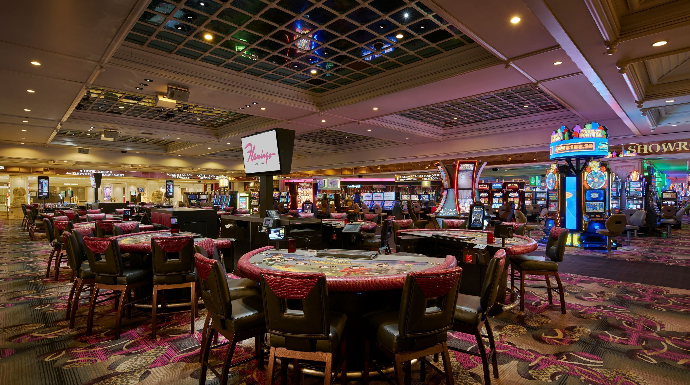A casino with multiple gaming tables and chairs, a large screen displaying 'Flamingo Las Vegas', and various slot machines.