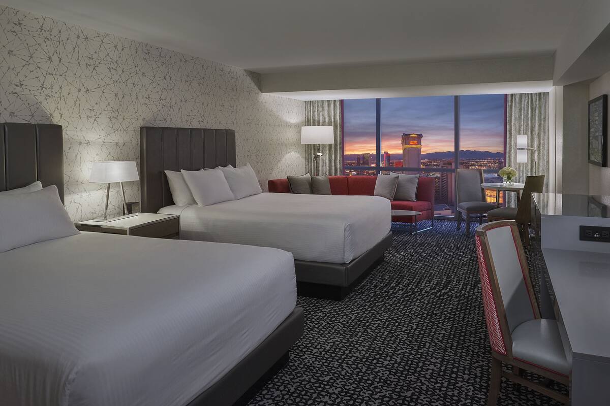 A hotel room with two beds, a desk, a chair, and a view of the cityscape at dusk.