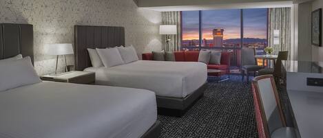 A hotel room with two beds, a desk, a chair, and a view of the cityscape at dusk.