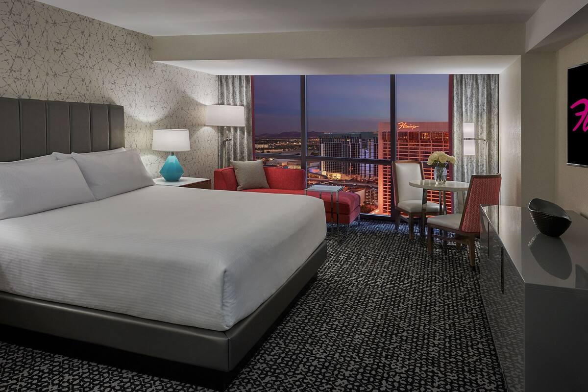 A hotel room with a large bed, a desk with a chair, a small table with a vase, and a view of the city at night.