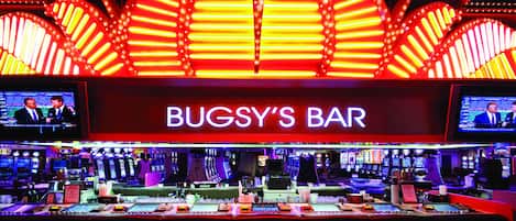 Un bar avec une enseigne au néon sur laquelle on peut lire « BUGSY’S BAR » et plusieurs écrans diffusant différents contenus.
