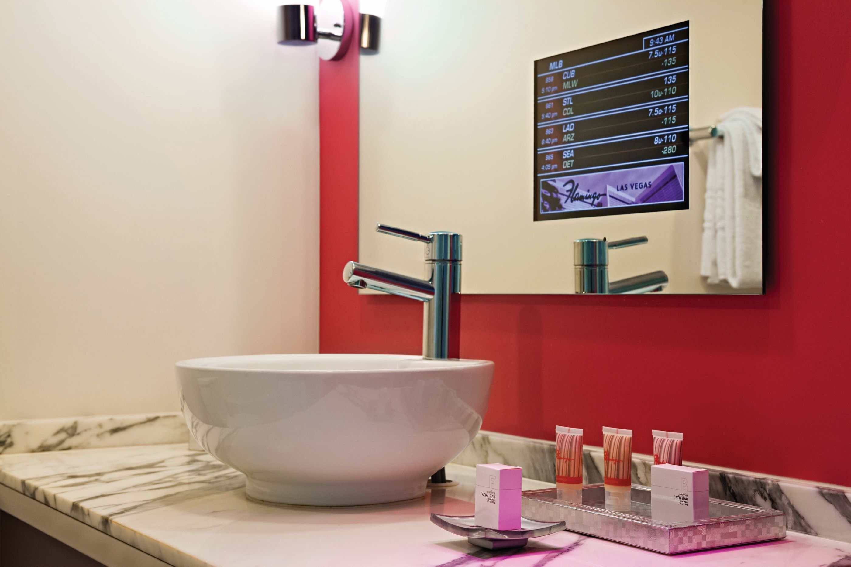 A bathroom with a large white sink, a mirror, and a digital display showing sports scores.
