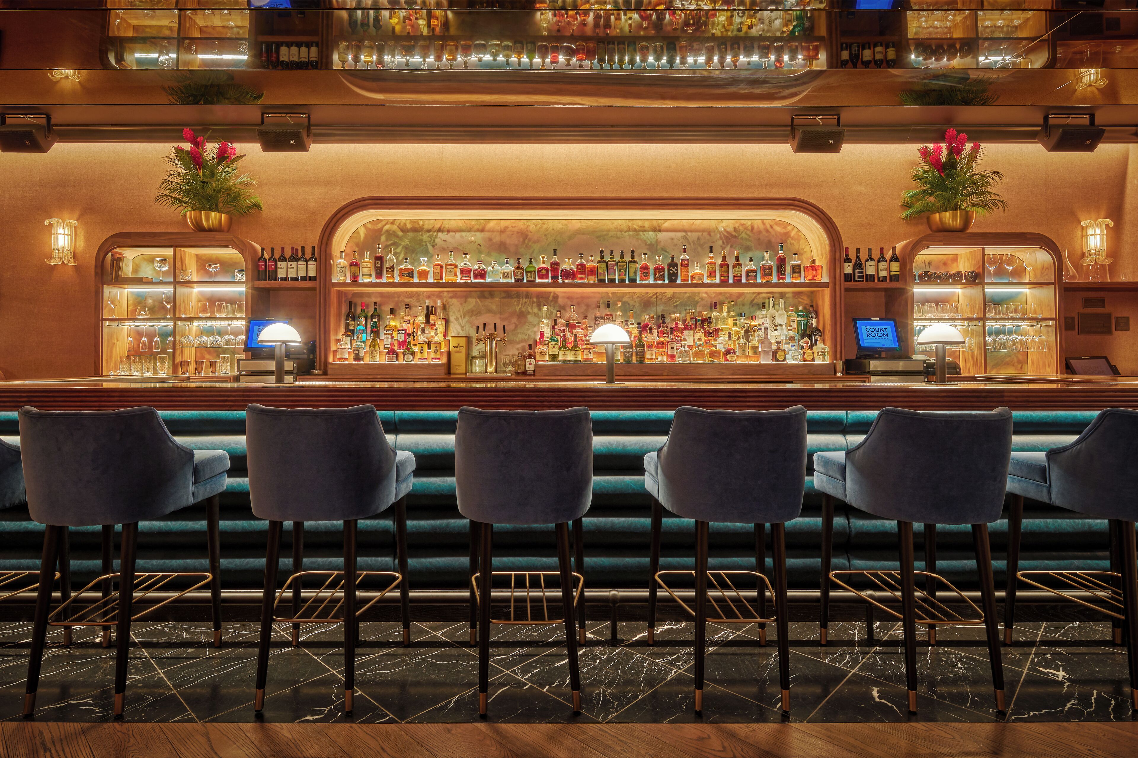 Una zona de bar con una barra larga, varias estanterías llenas de botellas y tres taburetes.