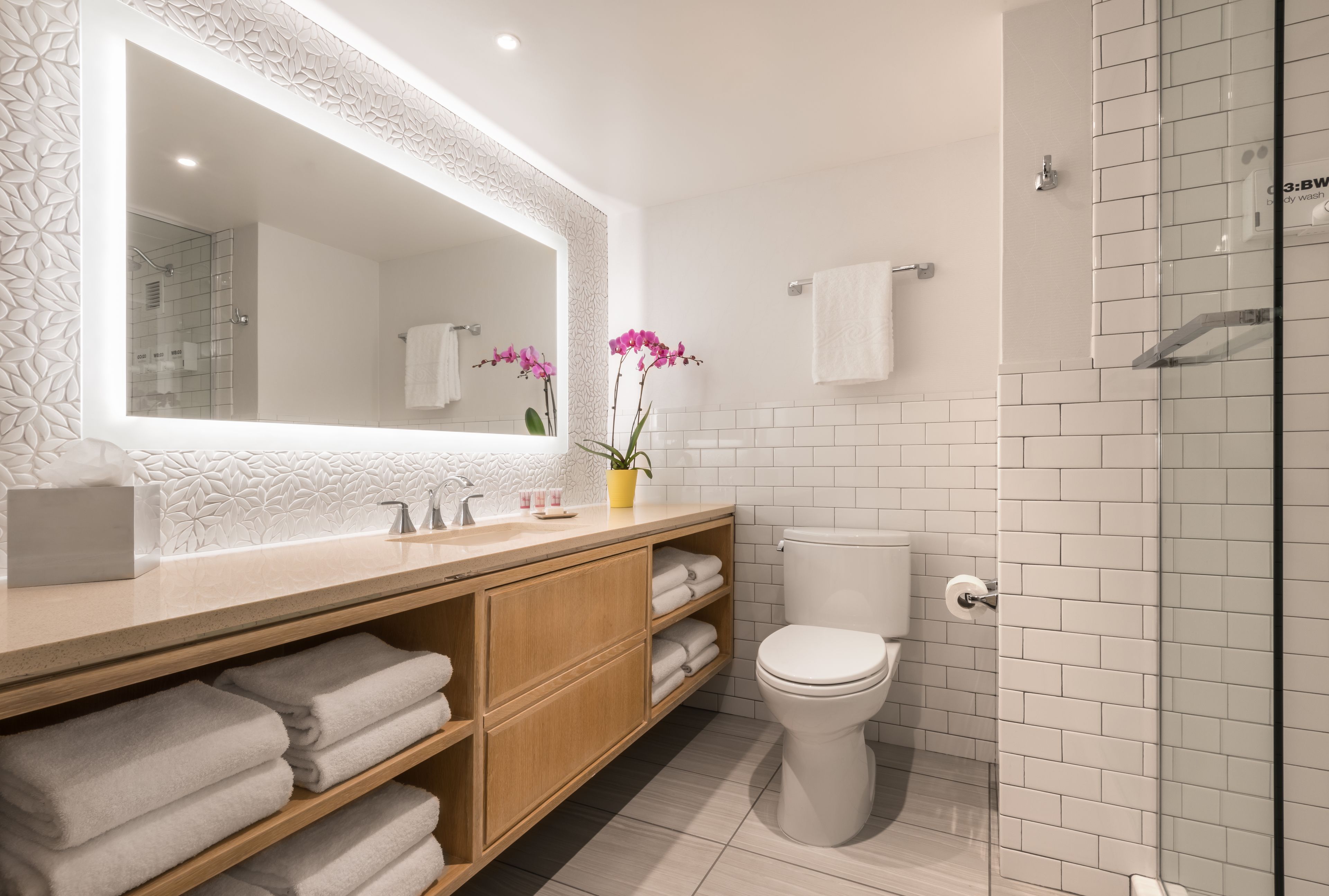 Een moderne badkamer met een grote spiegel, witte tegels, een houten wastafelmeubel, een toilet en een douchecabine met glazen wanden.