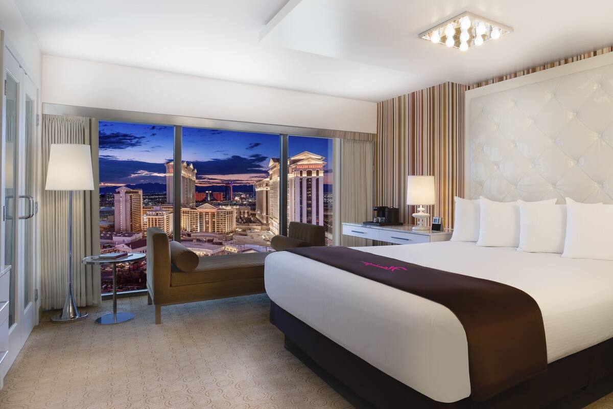 A hotel room with a large bed, a desk, a chair, and a view of a cityscape.
