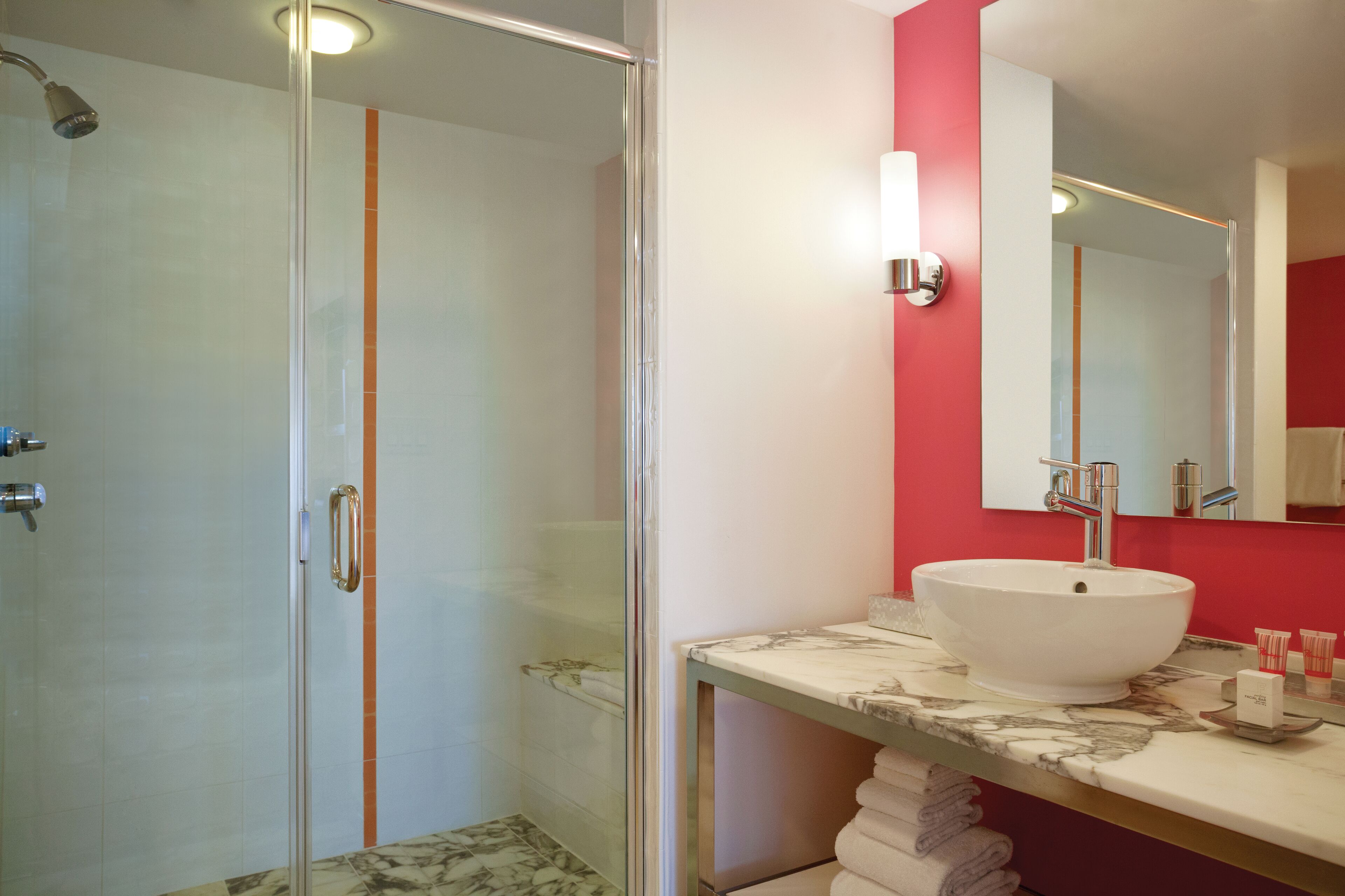 A modern bathroom with a glass shower enclosure, a white sink with a marble countertop, and a red accent wall.