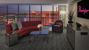 A modern hotel room with a city view, a red sofa, a grey armchair, a coffee table, and a TV displaying 'Flamingo'.