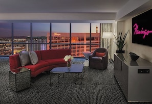 A modern hotel room with a city view, a red sofa, a grey armchair, a coffee table, and a TV displaying 'Flamingo'.