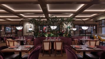Ein Restaurant mit Holztischen und violett gepolsterten Stühlen, umgeben von Topfpflanzen und gerahmten Bildern an den Wänden.