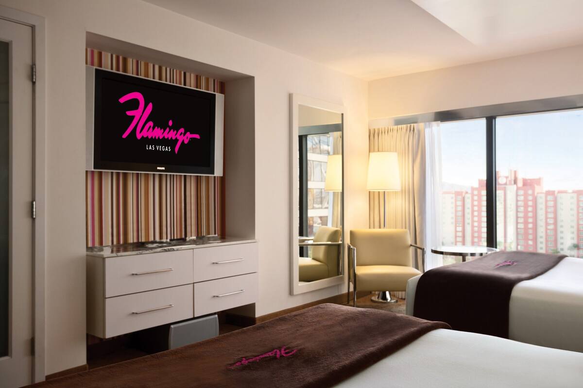 A hotel room with a flat-screen TV on the wall, a desk with drawers, a chair, a bed, and a window with a city view.