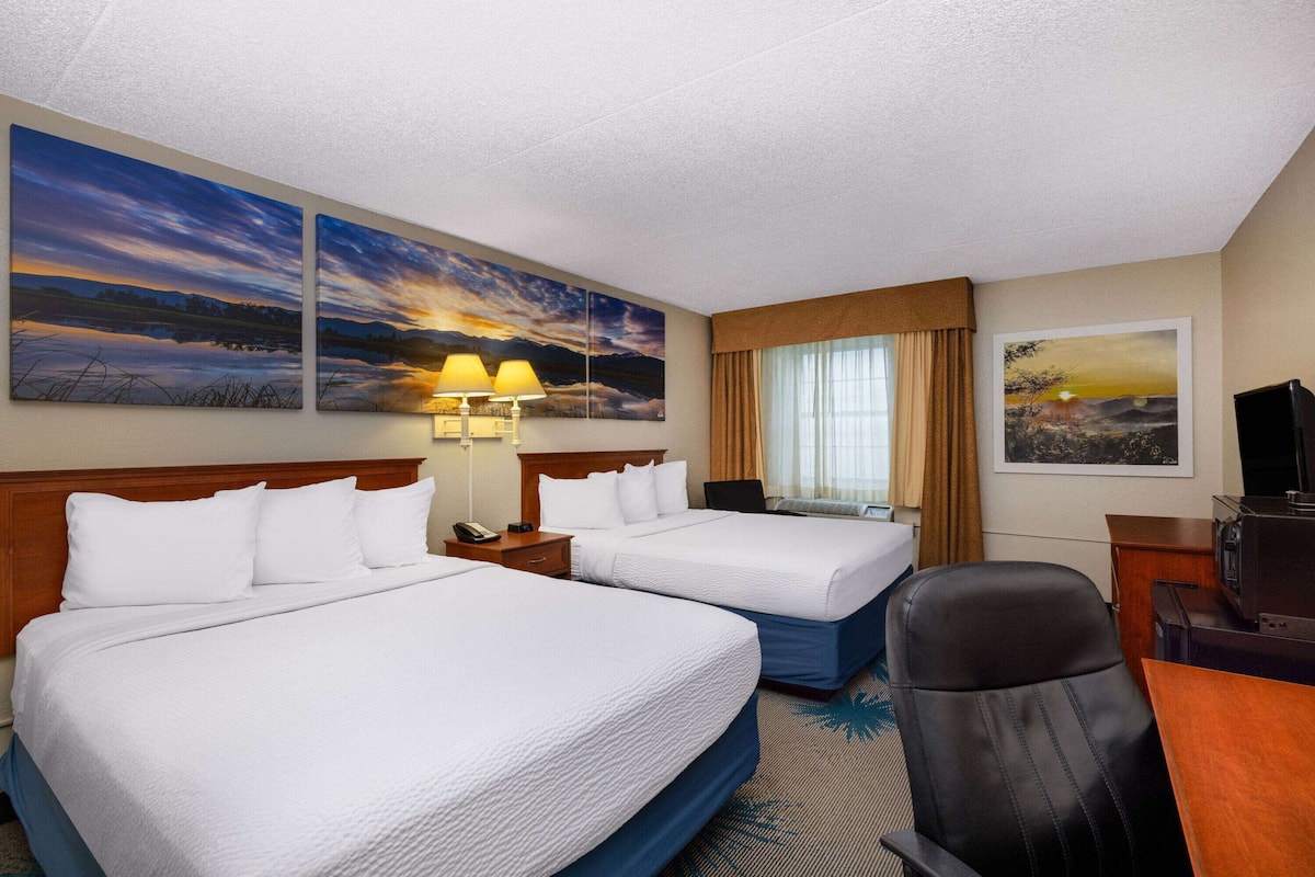 A hotel room with two beds, a desk, a chair, a TV, and artwork on the walls.