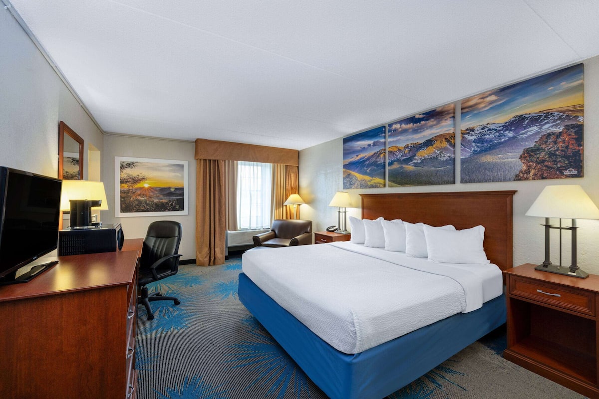 A hotel room with a large bed, a desk with a TV, two chairs, a desk lamp, and a painting on the wall.