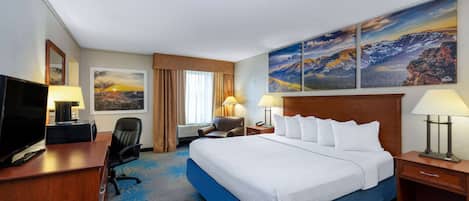 A hotel room with a large bed, a desk with a TV, two chairs, a desk lamp, and a painting on the wall.