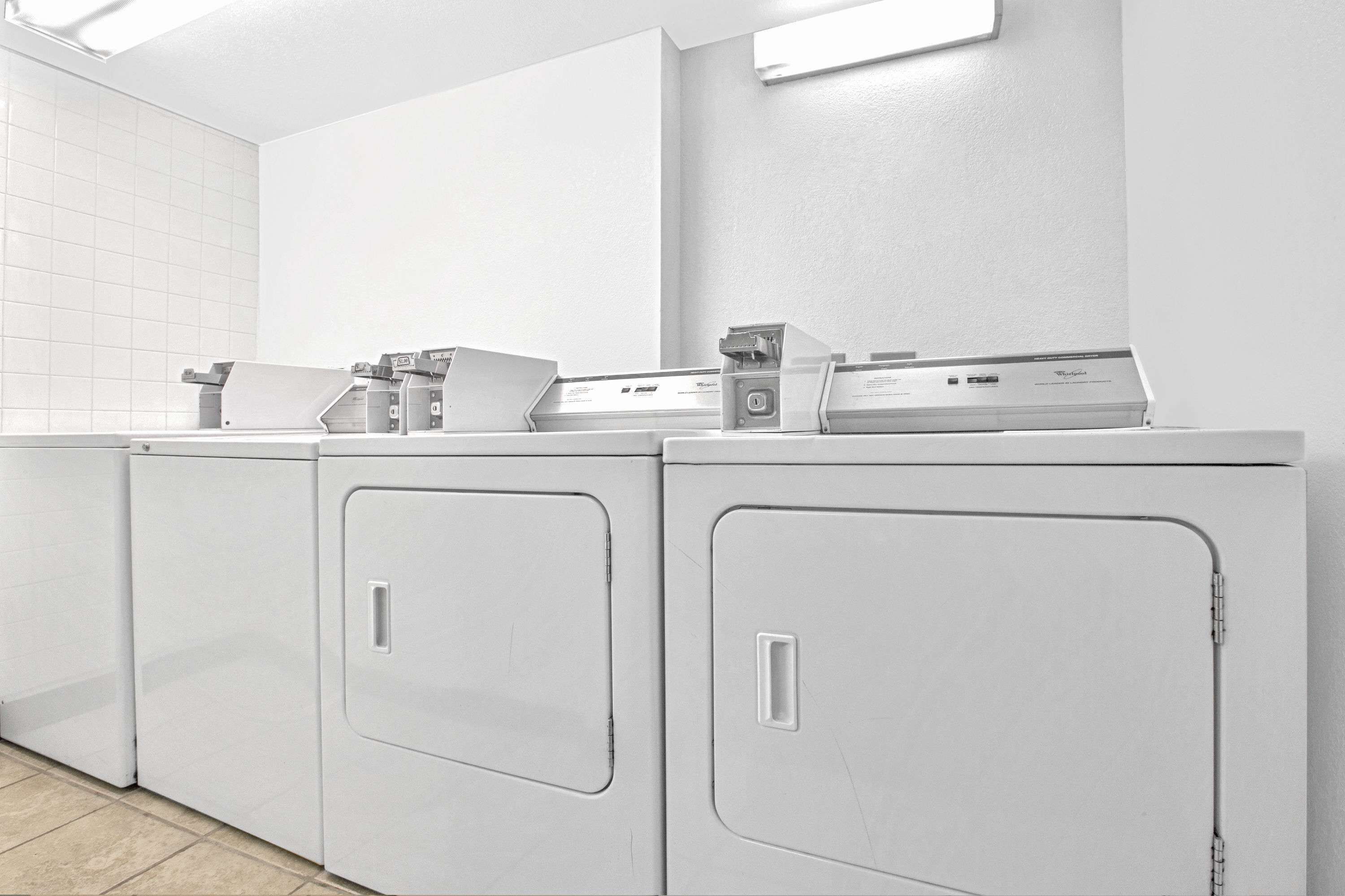 A row of white front-loading washing machines in a laundry room with a tiled wall and a light fixture above.