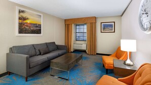 A hotel room with a gray sofa, a small table, an orange armchair, a blue patterned carpet, a painting of a sunset, a window with curtains, and a wall clock. - Days Inn by Wyndham Casper (Casper)