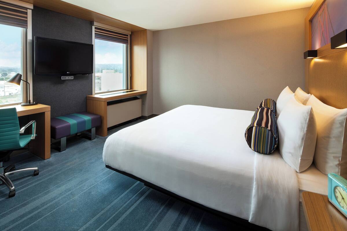 A hotel room with a large bed, a desk with a chair, a TV mounted on the wall, and a window with a view.