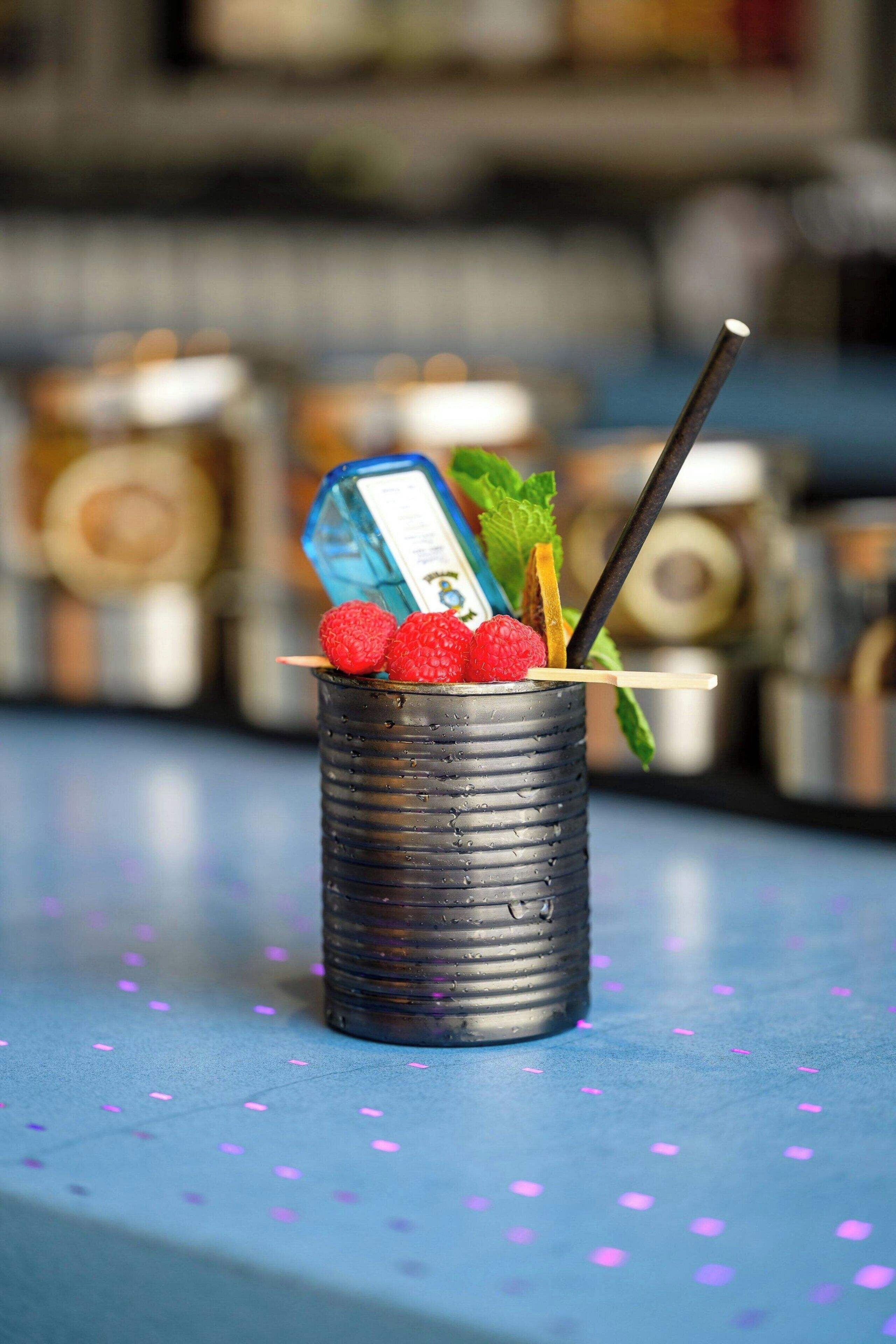 A cocktail in a textured glass with raspberries and mint leaves, placed on a blue surface with scattered dots.