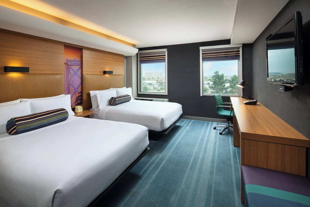 A hotel room with two beds, a desk, a TV, and a city view.