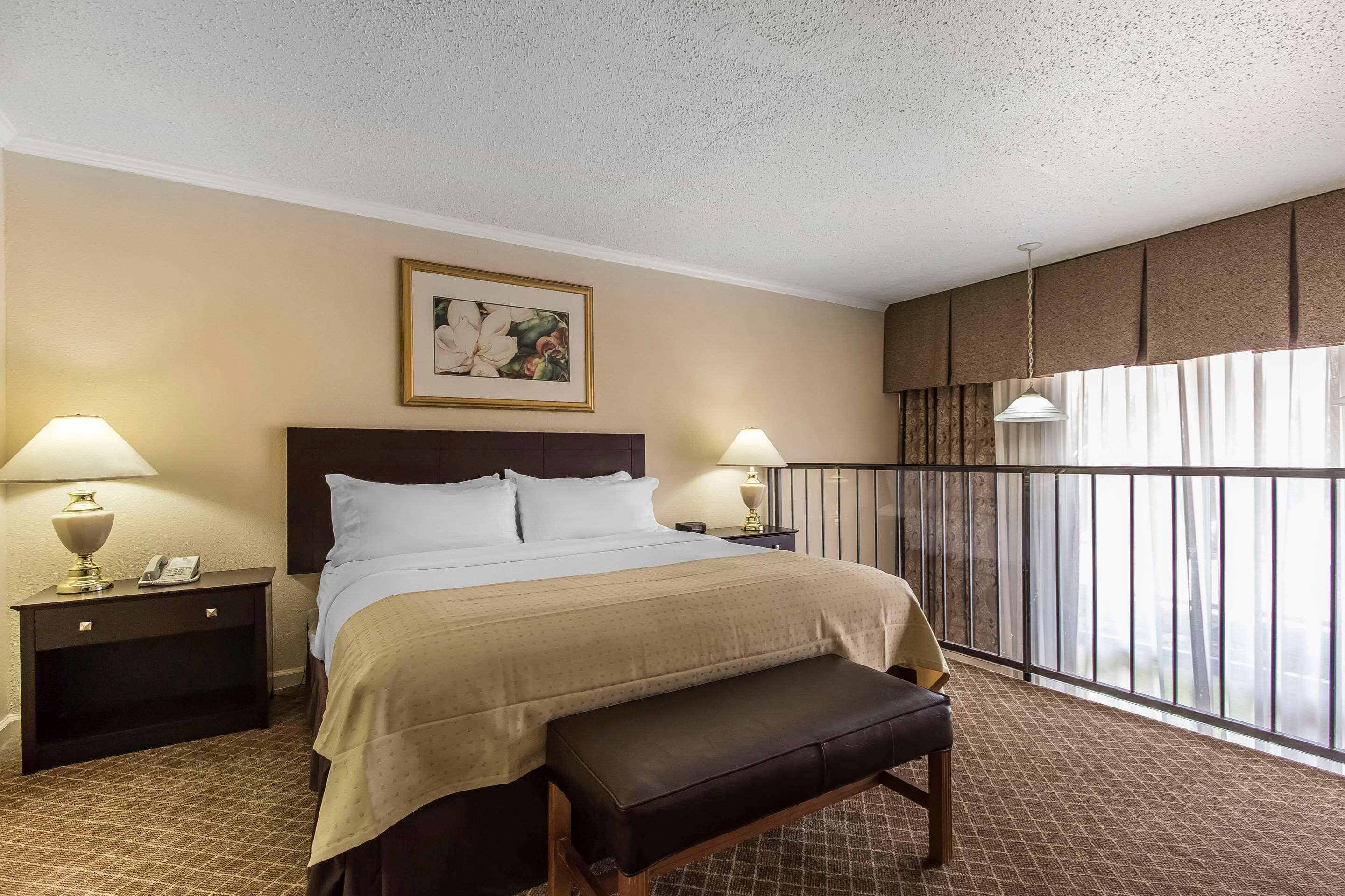 Habitación de hotel con una cama grande, dos mesitas de noche con lámparas, un banco, un cuadro en la pared y un balcón con vistas.