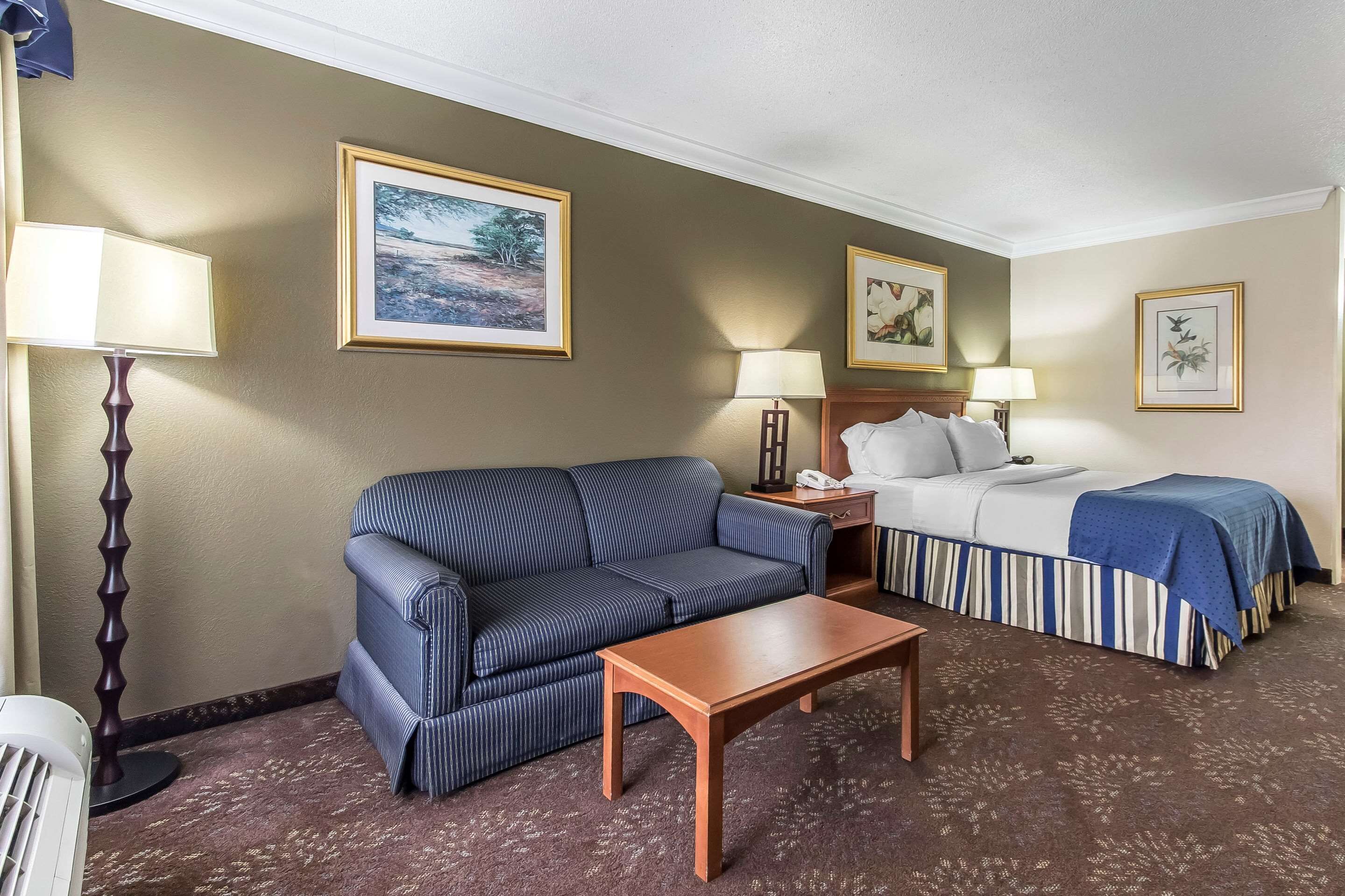 A hotel room with a striped sofa, a wooden coffee table, a bed with a striped bedspread, two bedside lamps, and framed artwork on the walls.