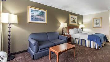 Habitación de hotel con un sofá a rayas, una mesa de centro de madera, una cama con colcha a rayas, dos lámparas de noche y obras de arte enmarcadas en las paredes.