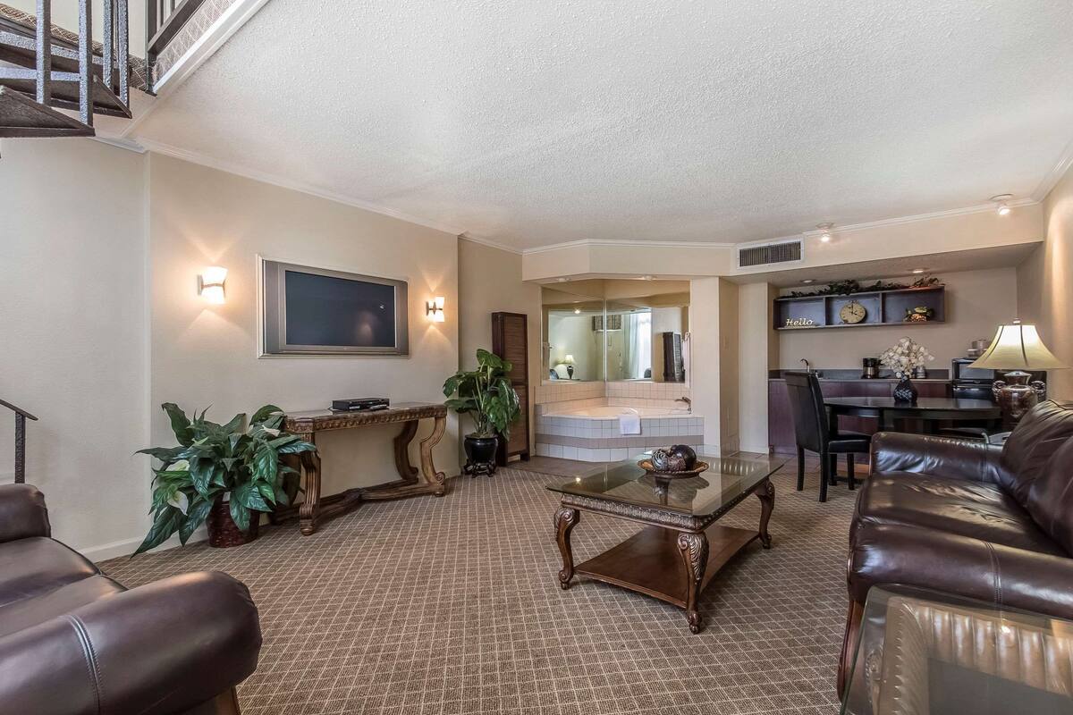 A living room with a sofa, coffee table, and a TV mounted on the wall. There is a staircase leading to an upper level, and a dining area with chairs and a table.