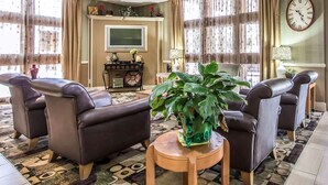 A living room with leather chairs, a round wooden table with a plant, a television, and a clock on the wall. - Clarion Inn Morgan City (Morgan City)