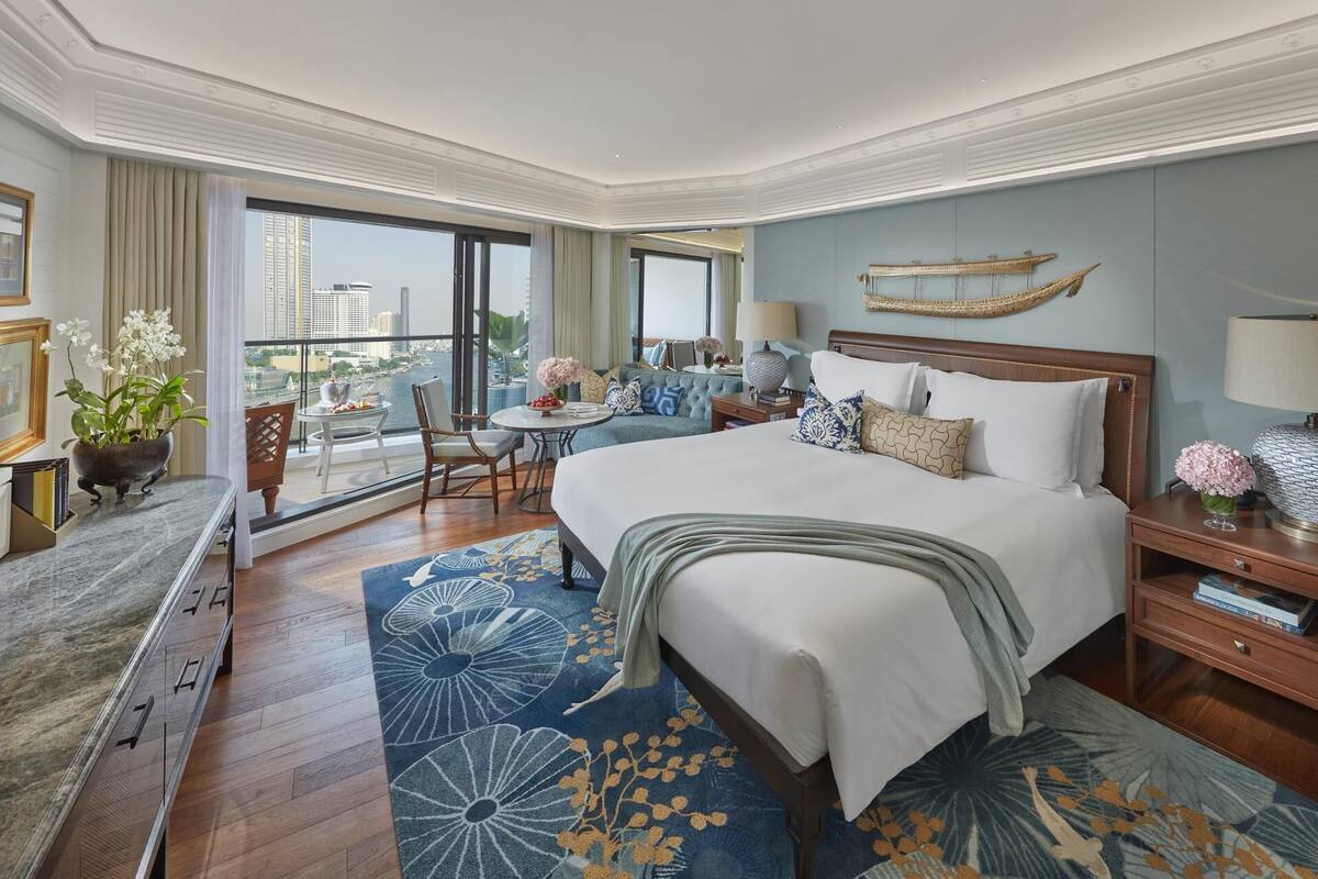 Habitación de hotel con una cama grande, un balcón con vistas a la ciudad, una mesa redonda y un rincón de descanso con una silla y una mesita.