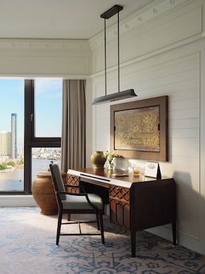 A modern hotel room with a large window offering a city view, a desk with a vase of flowers, and a decorative wall piece.