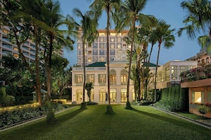 A large hotel with a symmetrical facade, multiple balconies, and a well-maintained garden with palm trees and hedges.