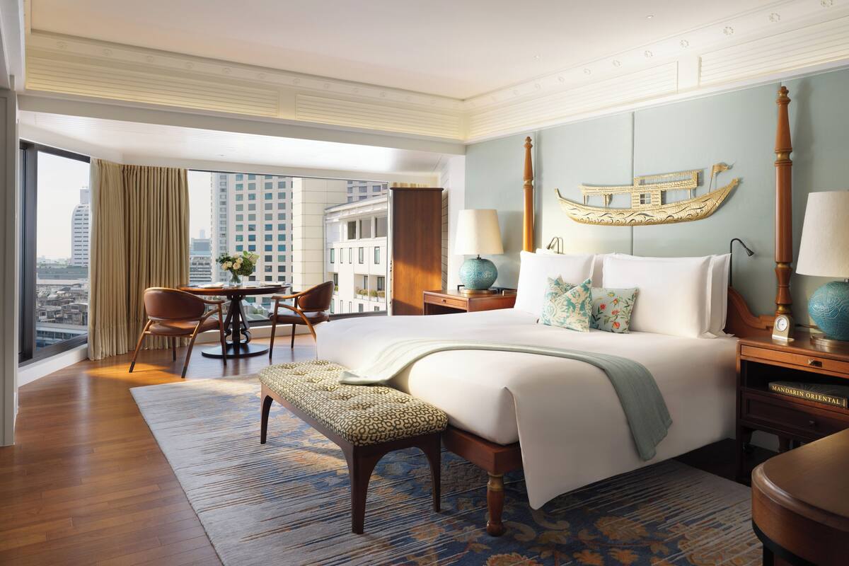 Una habitación de hotel amplia con una cama grande, una zona de estar con una mesa y sillas, vistas de la ciudad a través de amplios ventanales y un elemento decorativo en la pared.