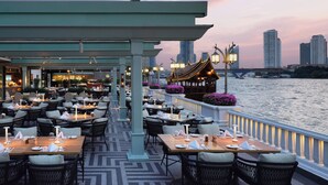 An outdoor dining area with tables and chairs, a covered section, and a view of the river and city skyline. - Mandarin Oriental, Bangkok (Bangkok)
