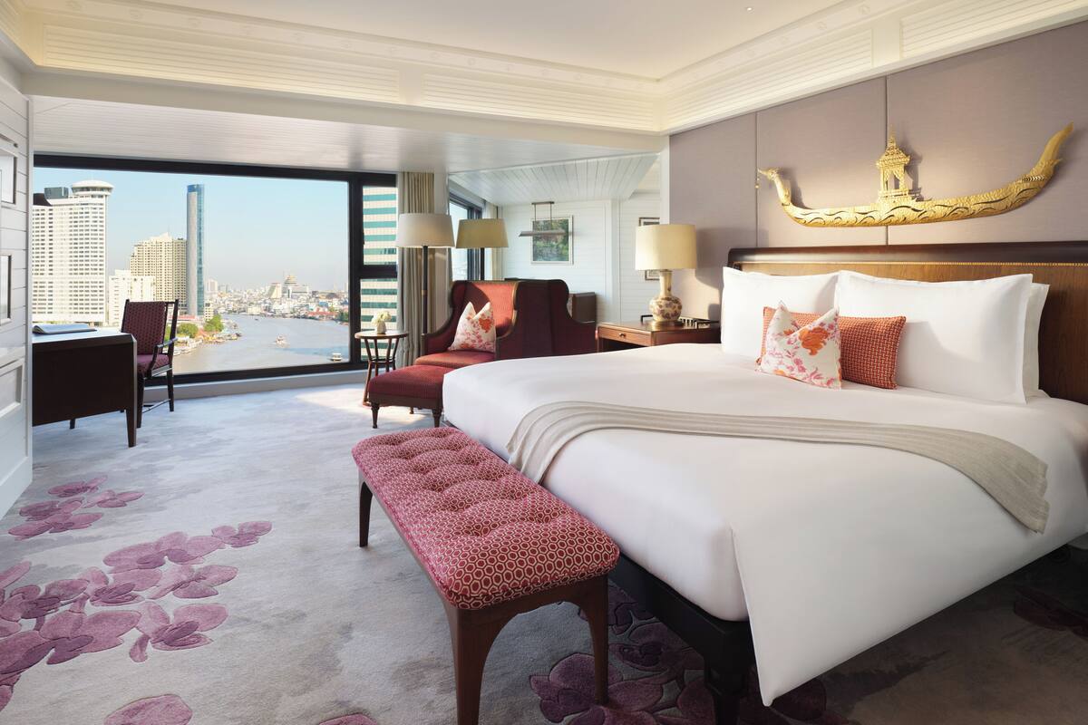 Habitación de hotel con una cama grande, un banco, un escritorio y una silla. Se ve un paisaje urbano a través de grandes ventanales.