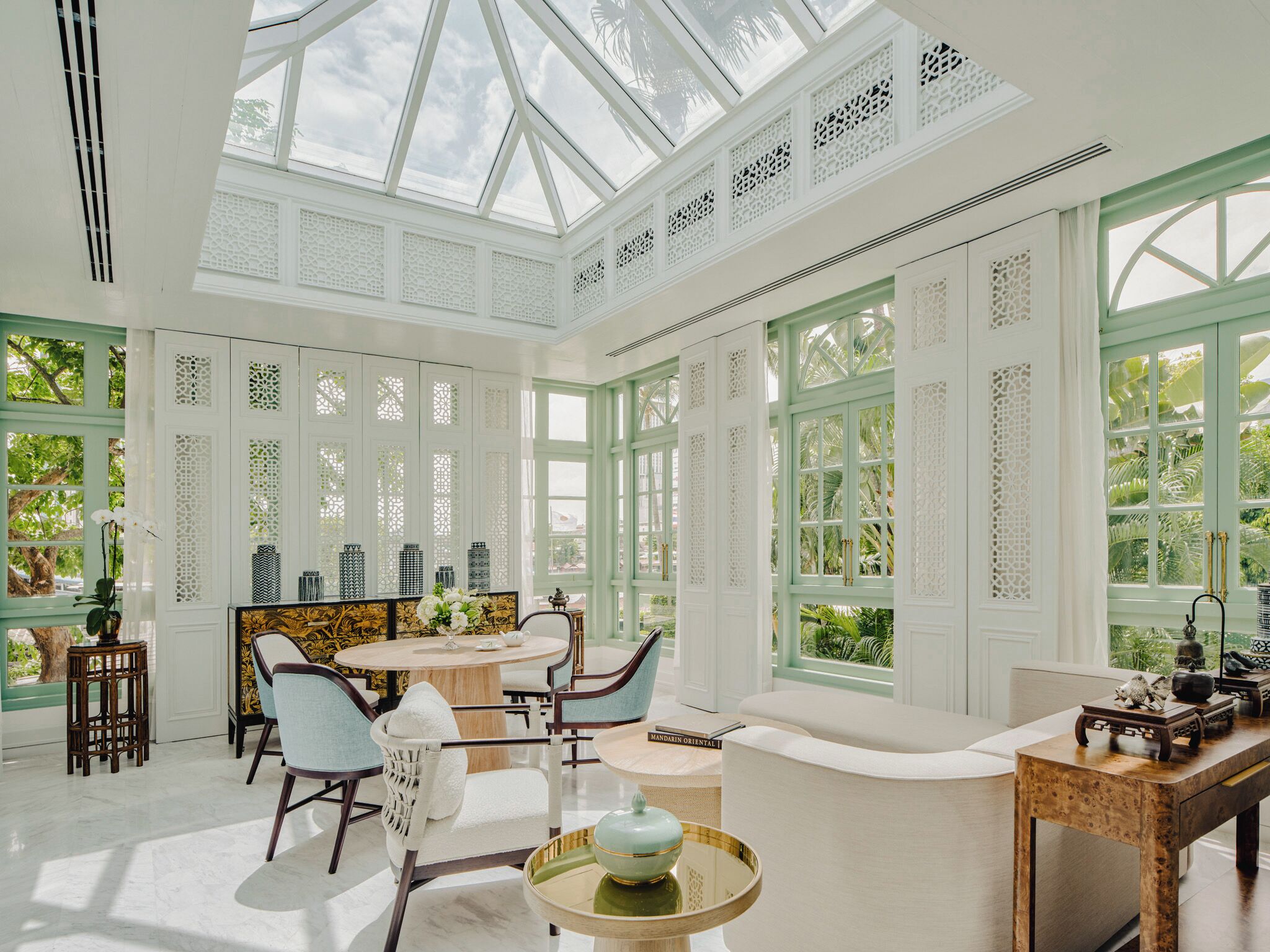 A spacious room with a high ceiling and large windows, featuring a dining area with a round table and chairs, a seating area with a sofa and armchairs, and a small side table with a decorative bowl.