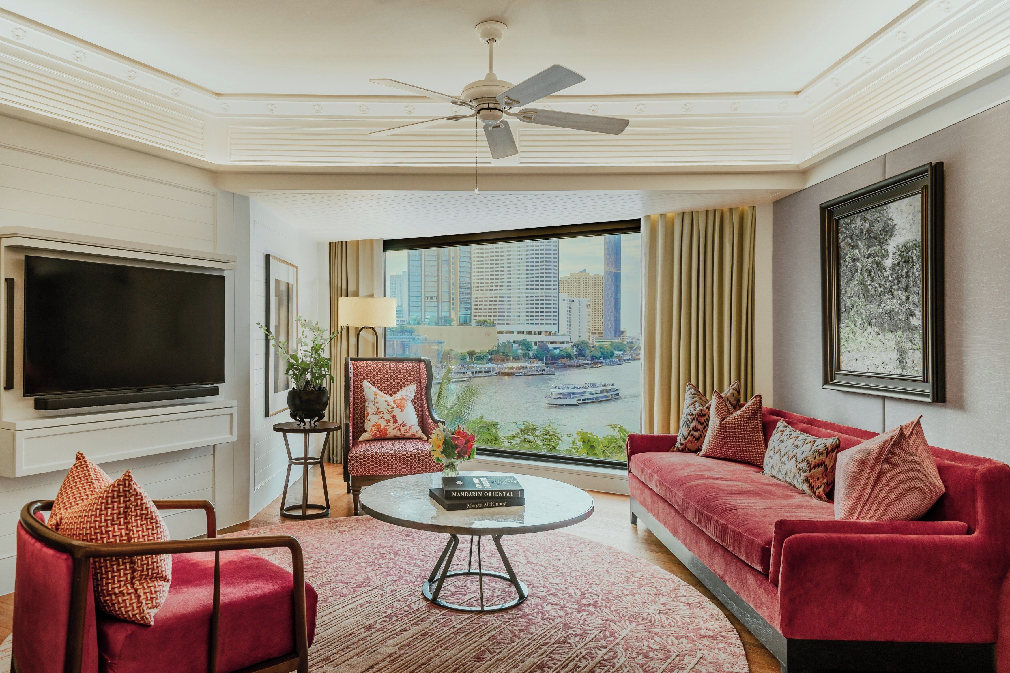 A modern living room with a flat-screen TV, a red sofa, a red armchair, a coffee table, and a view of a cityscape through large windows.