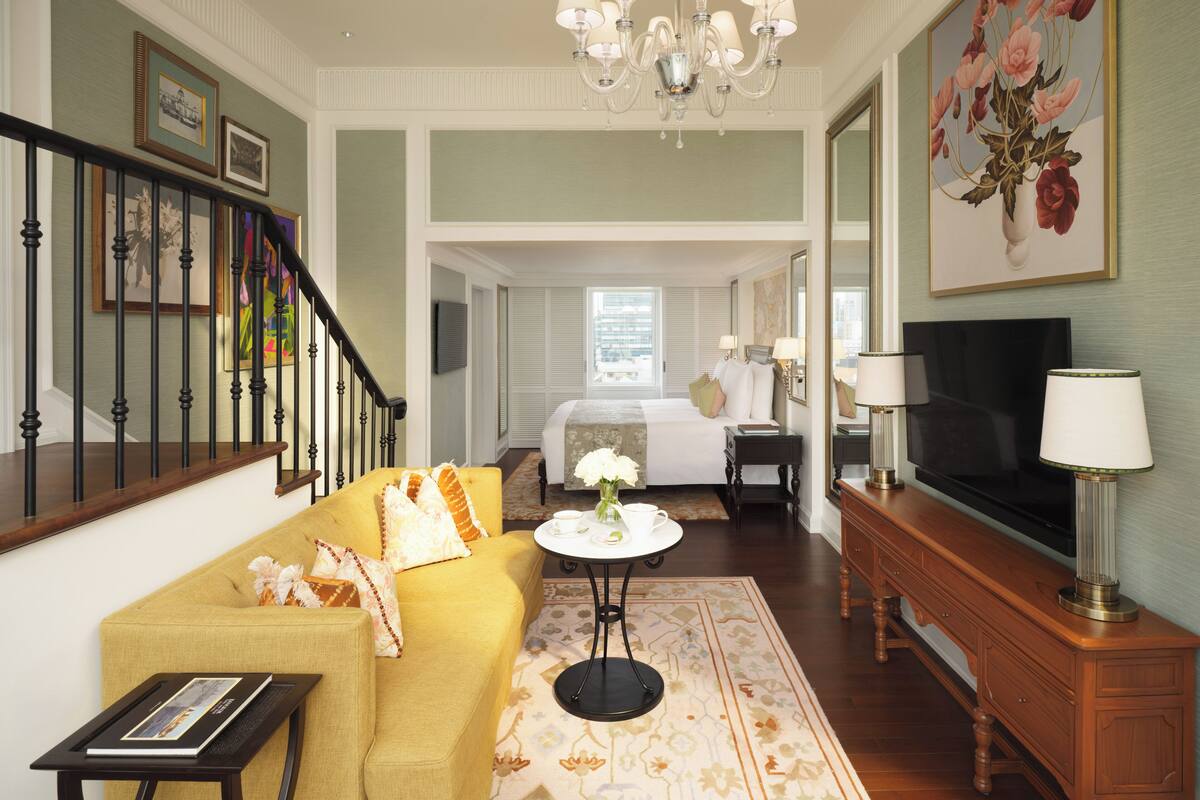 Habitación de hotel con sofá amarillo, una mesita de centro, televisor, candelabro y un cuadro de flores.