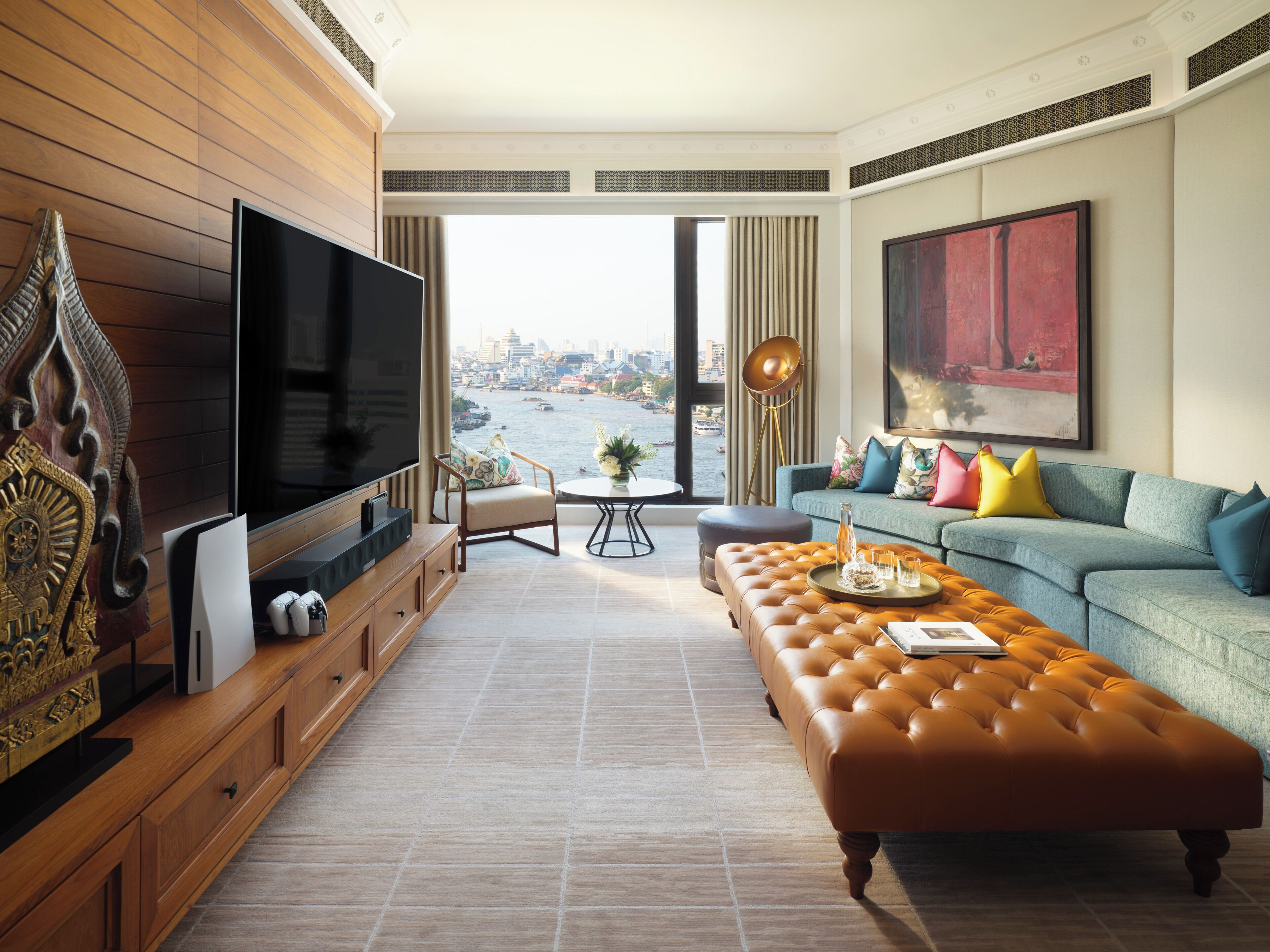 A modern living room with a large flat-screen TV, a sofa with colorful cushions, a leather ottoman, and a view of a cityscape through a large window.