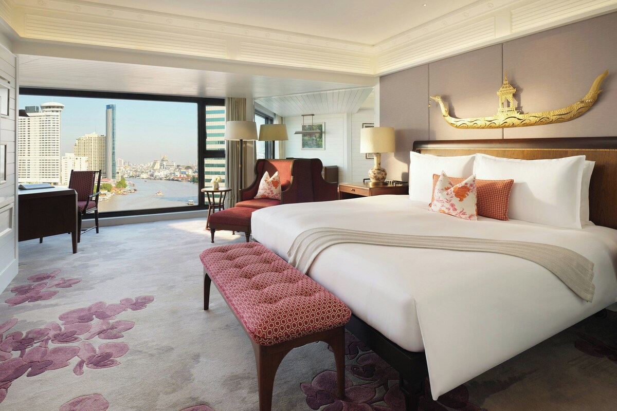 Habitación de hotel con una cama grande, un banco, un escritorio y una silla. Se ve una vista de la ciudad a través de la ventana.