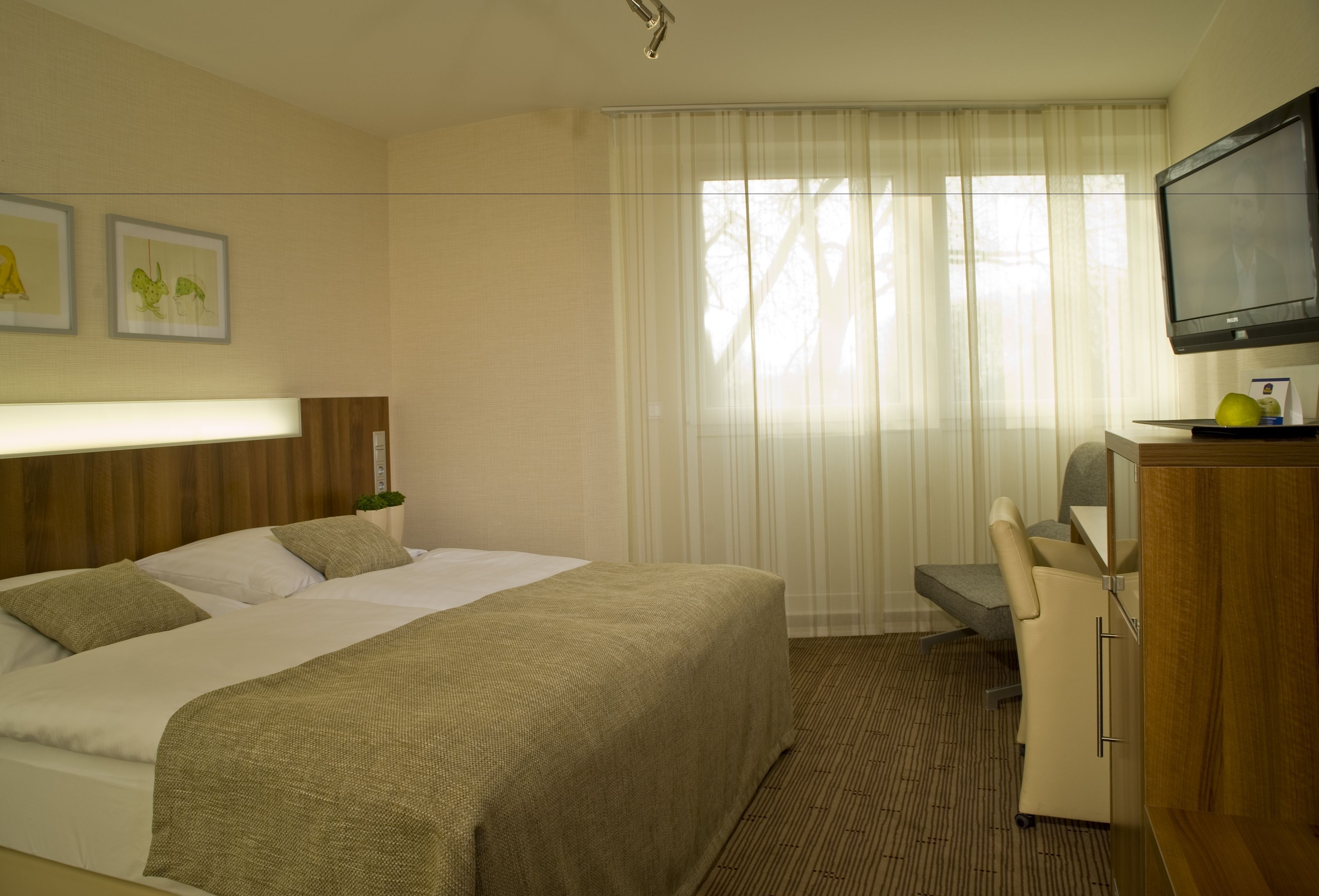 A hotel room with a large bed, a desk, a chair, a television, and a window with sheer curtains.