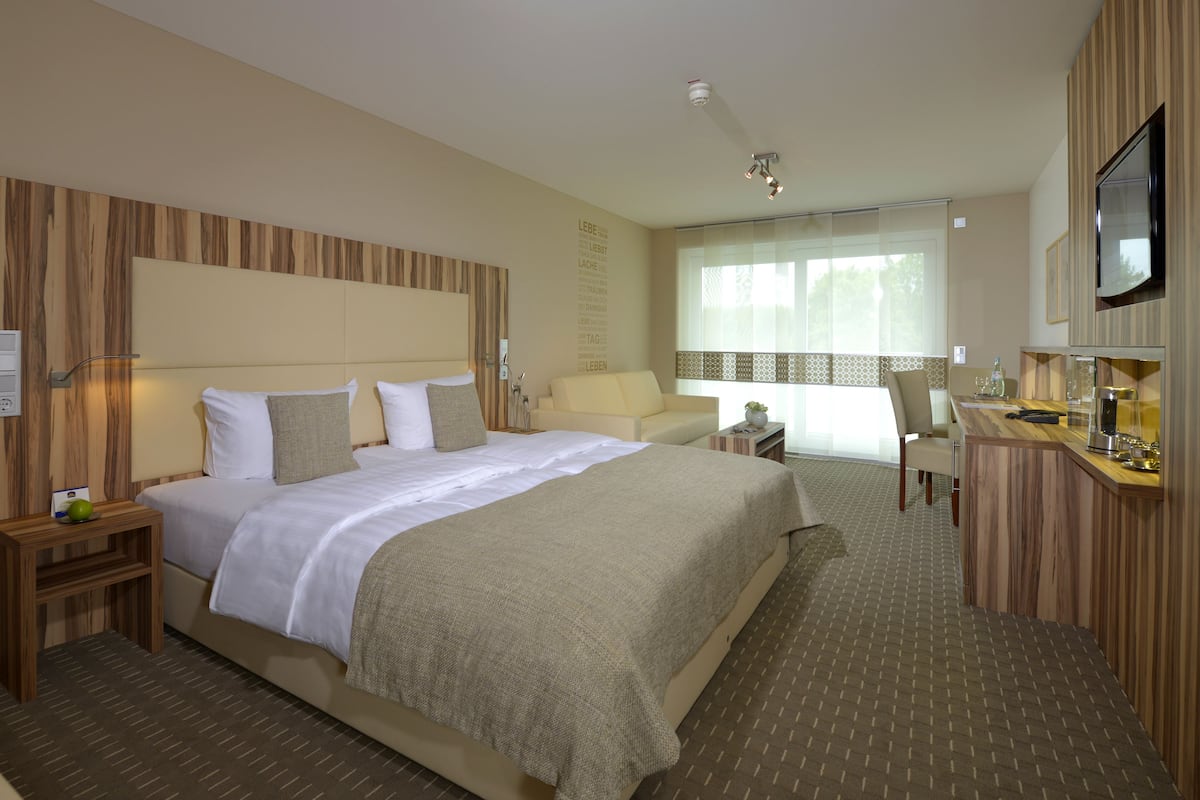 A hotel room with a large bed, bedside tables, a desk with a chair, a TV, and a window with curtains.
